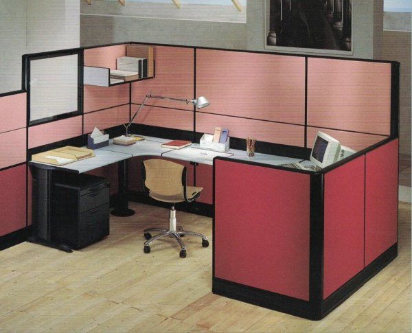 Un cubicolo d'ufficio a forma di L con divisori in tessuto rosa chiaro e rosso, una scrivania bianca, una sedia girevole e attrezzature informatiche.
