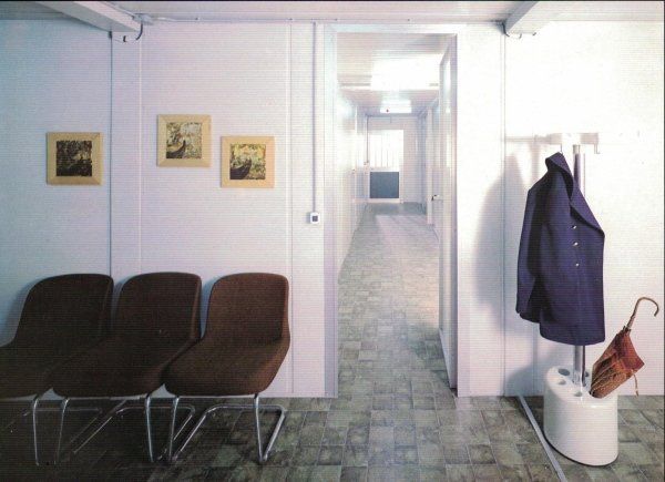 Sala d'attesa con tre sedie marroni, un quadro incorniciato su una parete bianca e un appendiabiti con giacca e ombrello.