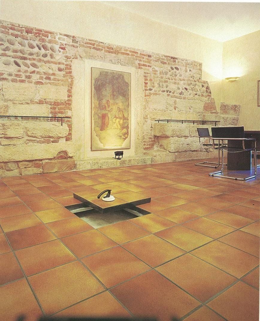 Una stanza interna con pareti rustiche in mattoni a vista, un quadro incorniciato, pavimento in piastrelle di terracotta e una piccola botola sul pavimento.