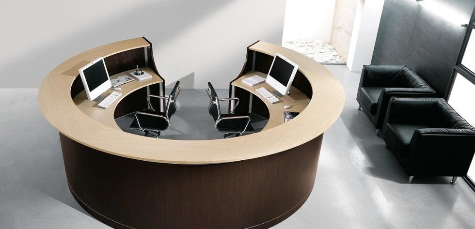 Un bancone reception curvo in legno scuro per due persone con ripiani chiari, due computer e sedie nere nelle vicinanze.