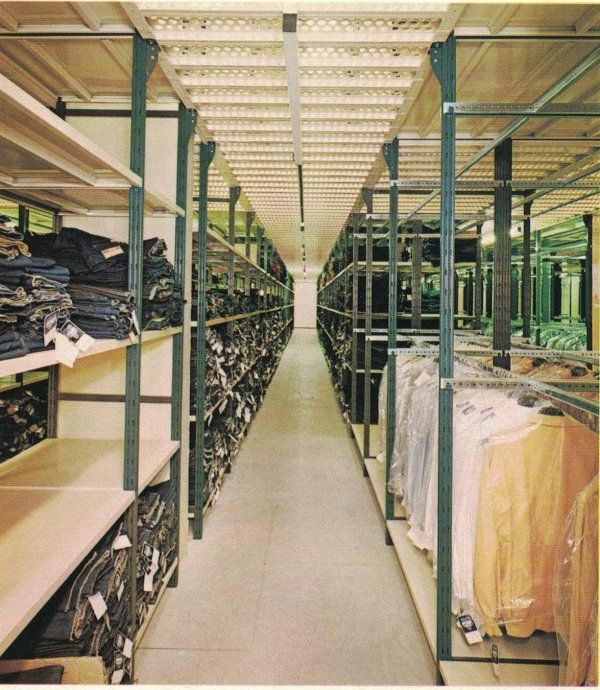 Uno stretto corridoio di un magazzino, caratterizzato da alte scaffalature metalliche piene di jeans piegati e indumenti appesi su entrambi i lati.