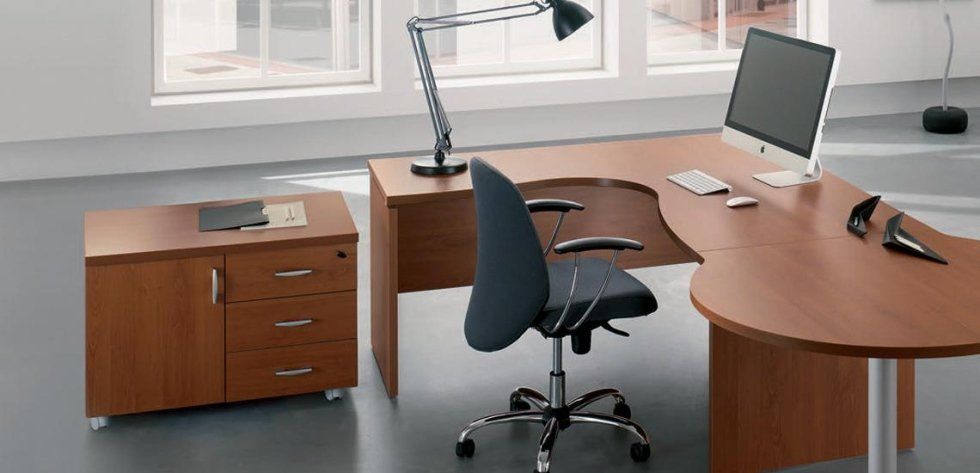 Una scrivania angolare in legno con un mobiletto a cassetti abbinato, una sedia da ufficio girevole, un computer e una lampada da scrivania.