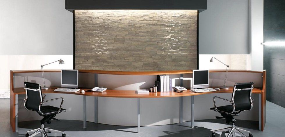 Una moderna reception in legno dalle linee curve, con due postazioni computer e sedie da ufficio nere, addossata a una parete in pietra.