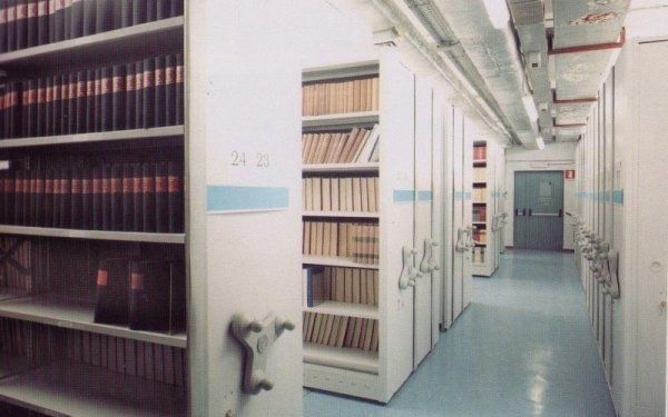 Scaffalature ad alta densità piene di libri in un archivio bibliotecario con pavimento azzurro e meccanismi di scorrimento in metallo.