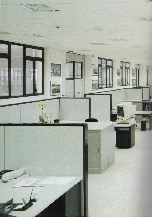Ufficio vuoto con file di cubicoli e scrivanie grigie, grandi finestre lungo la parete e luci fluorescenti a soffitto.