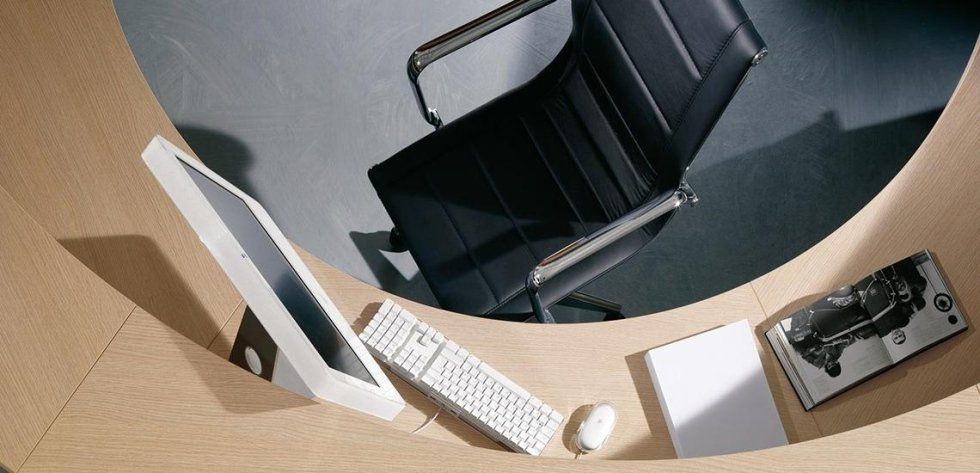 Una scrivania curva in legno chiaro con computer, tastiera, mouse, notebook e telefono, accanto a una sedia da ufficio nera.