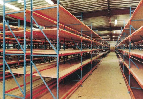 Magazzino vuoto con file di scaffalature industriali alte, con struttura blu, travi arancioni e ripiani in legno.