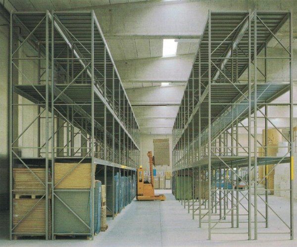 Un carrello elevatore arancione nel corridoio di un magazzino, tra file di alte scaffalature metalliche industriali di colore grigio.