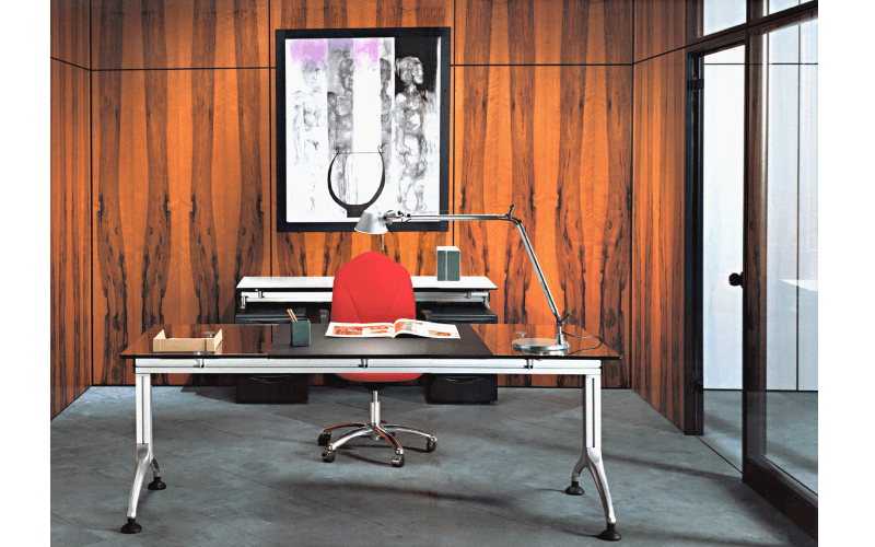 Ufficio moderno con scrivania con piano in vetro scuro, sedia rosso acceso e un quadro astratto incorniciato su una parete rivestita in legno.