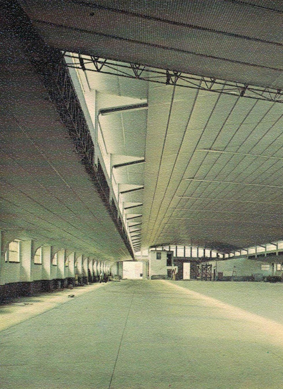 Interno di un ampio capannone industriale vuoto con soffitto alto, travi strutturali a vista e pavimento in cemento.