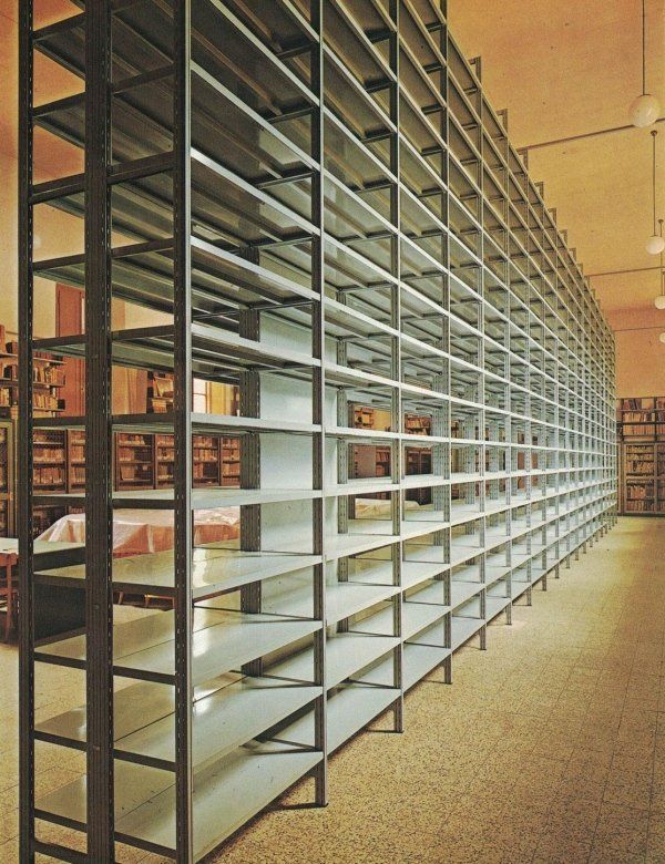 File di alte scaffalature metalliche da biblioteca che si estendono per tutta la lunghezza di una stanza con pavimento di colore chiaro.