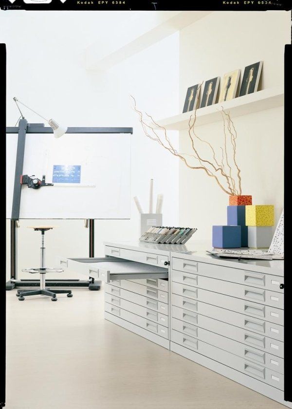 Uno studio luminoso e moderno, dotato di un grande tavolo da disegno bianco, uno sgabello e lunghe file di cassettiere in metallo bianco.