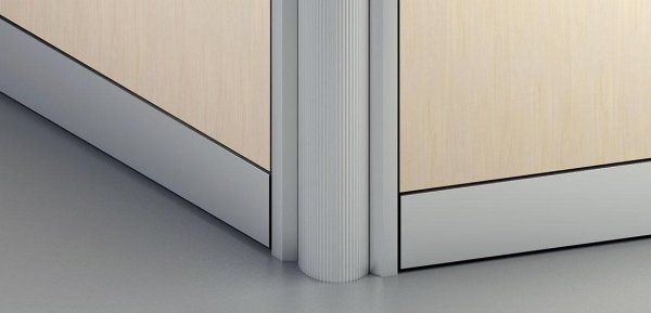 Primo piano di un montante angolare metallico arrotondato che collega due pareti divisorie per ufficio di colore chiaro con battiscopa argentati.