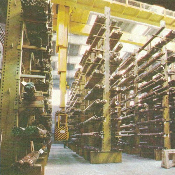 Un alto magazzino industriale con scaffalature metalliche su cui sono impilate lunghe barre, e una gru a ponte gialla nel corridoio.