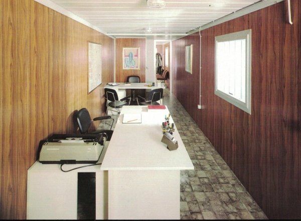 Un ufficio lungo e stretto all'interno di un container mobile, caratterizzato da pareti rivestite in legno, scrivanie bianche e pavimento decorato.