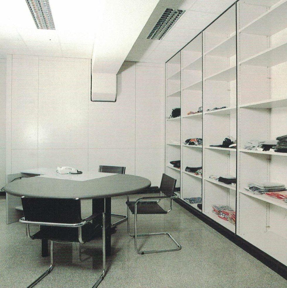 Ufficio minimalista con tavolo circolare grigio, sedie nere e scaffalature alte bianche lungo la parete.
