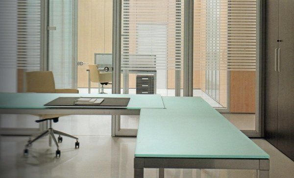 In un ufficio moderno con ampie finestre e arredi minimalisti, una scrivania a forma di L con piano in vetro verde chiaro è collocata.