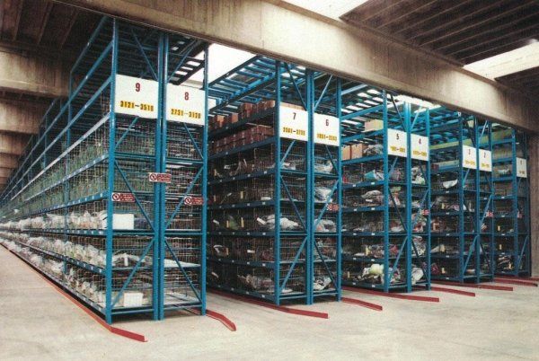 Scaffalature industriali in metallo blu disposte in file con cartelli numerati in un magazzino con soffitto in cemento.