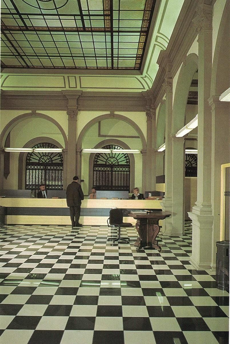 Vista interna di una hall con pavimento a scacchi, elementi architettonici ad arco e un bancone di servizio con personale.