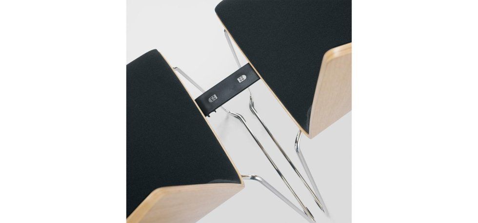 Due diffusori in legno con griglie in tessuto nero, collegati da un ponte centrale nero e sorretti da sottili supporti metallici.