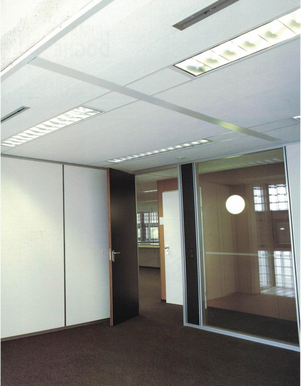 Interno di un ufficio caratterizzato da pareti divisorie bianche, moquette scura, una porta nera e un ufficio con pareti in vetro e una lampada rotonda.