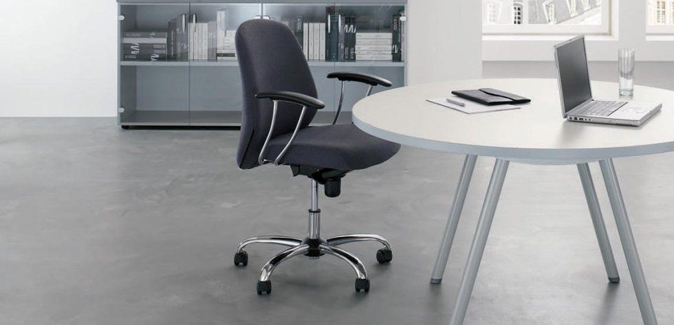 In un ufficio moderno con vetrina, una sedia da ufficio grigio scuro è posizionata accanto a un tavolo rotondo di colore chiaro su cui è appoggiato un computer portatile.