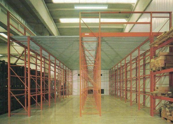 Interno di un magazzino industriale caratterizzato da un alto sistema di scaffalature a soppalco in metallo arancione con pavimento a rete.