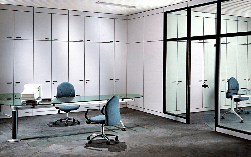 Un ufficio minimalista caratterizzato da una scrivania in vetro, due sedie color verde acqua e armadi bianchi a tutta parete su un pavimento grigio scuro.