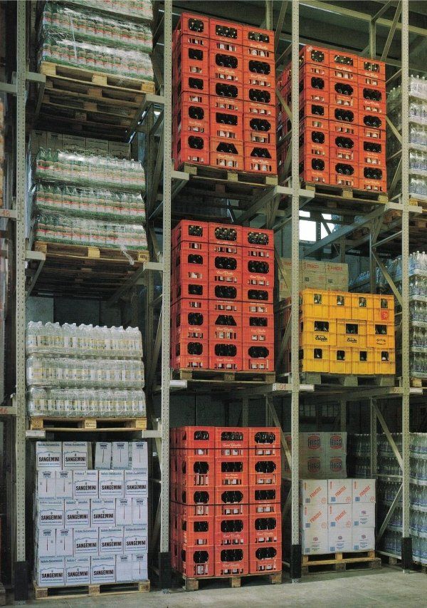 Sistema di scaffalature industriali per magazzino, pieno di pallet contenenti casse di bevande rosse e gialle impilate e scatole bianche.
