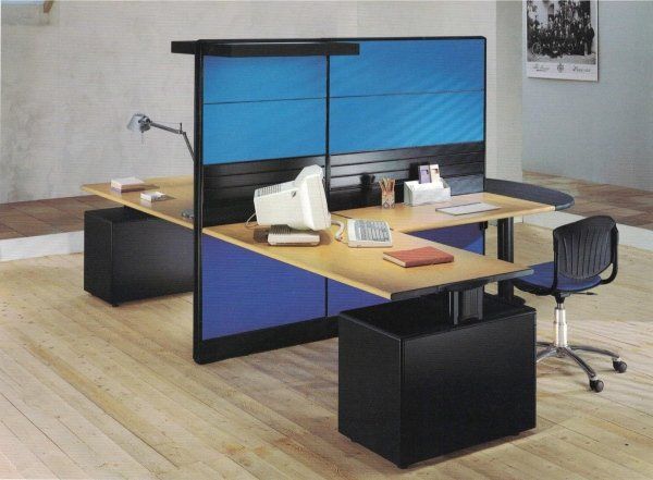 Postazione di lavoro per ufficio a doppia faccia con scrivanie in legno chiaro, armadietti neri e divisori blu per garantire la privacy.