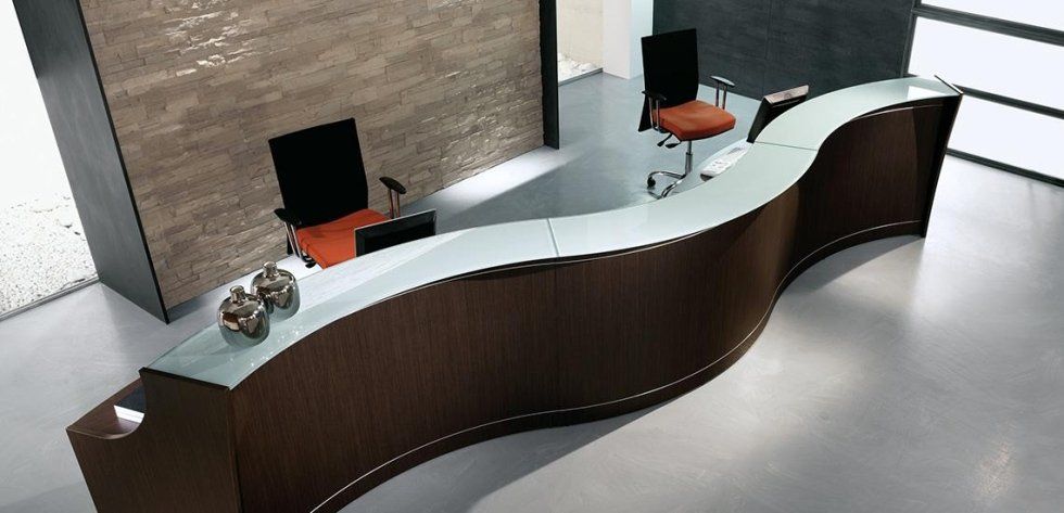 Un moderno bancone reception in legno dalle linee curve, con piano in vetro, e due sedie da ufficio, in un ambiente open space.