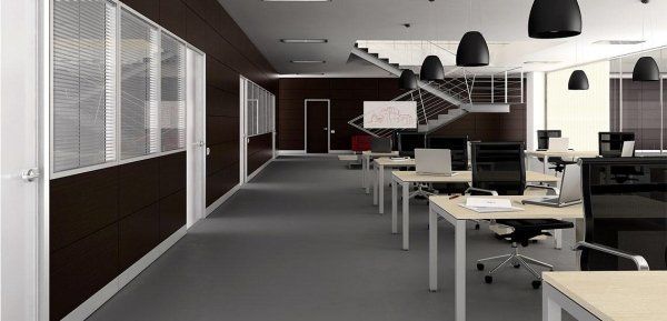 Un ufficio moderno con scrivanie vuote, file di lampade a sospensione nere, una parete scura a contrasto e una scala sullo sfondo.