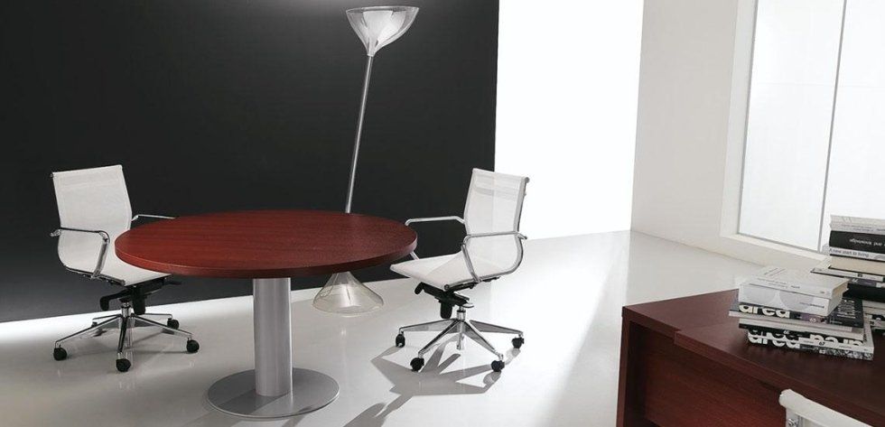 In un ufficio moderno e minimalista, attorno a un tavolo rotondo color mogano si trovano due sedie da ufficio bianche, illuminate da un'alta lampada da terra.