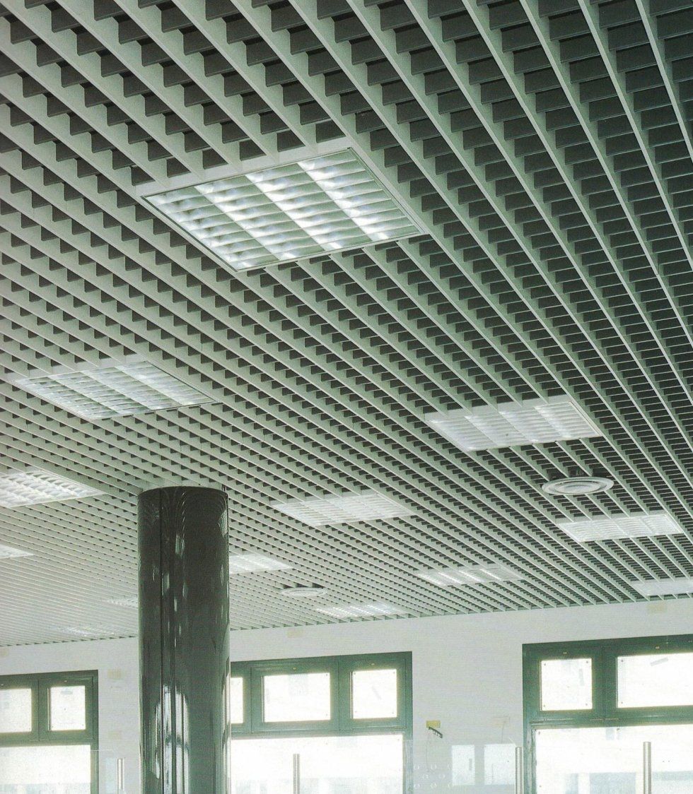 In una stanza con finestre, il soffitto è caratterizzato da una griglia bianca a celle aperte con faretti a incasso quadrati e una colonna verticale scura.