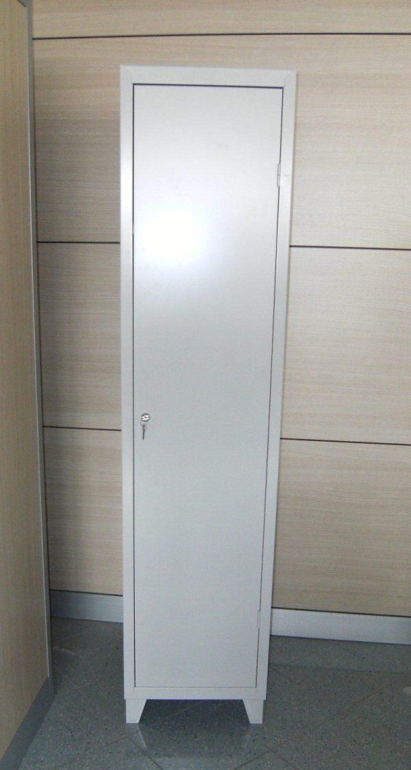 Un armadietto metallico bianco, alto e stretto, con serratura a chiave, appoggiato su gambe corte contro una parete rivestita di pannelli.