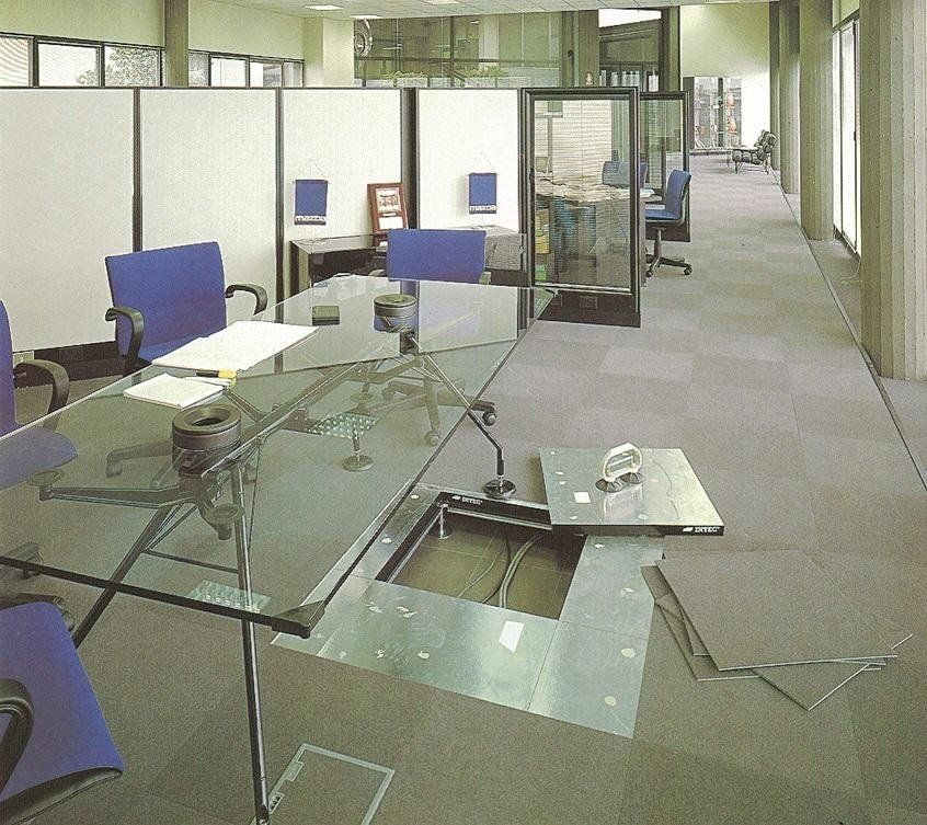Un pannello di accesso a pavimento a vista rivela il cablaggio all'interno di un ufficio con scrivania in vetro, sedie blu e pareti divisorie modulari.