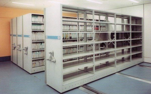 Scaffalature mobili ad alta densità per l'archiviazione di documenti, con manovelle manuali, in un ambiente ufficio bianco e asettico.