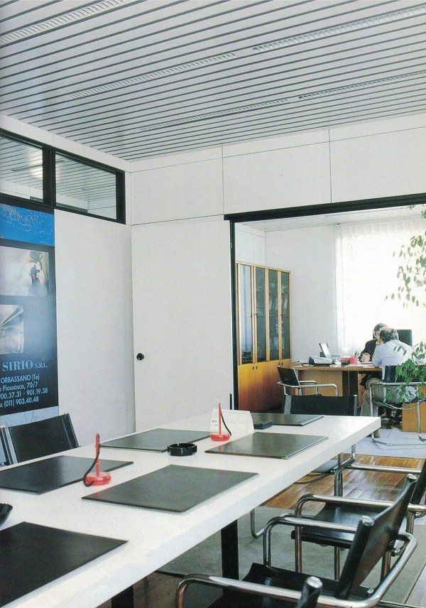 Una moderna sala conferenze con un lungo tavolo bianco e sedie nere, che si affaccia su un ufficio adiacente dove alcune persone stanno lavorando.