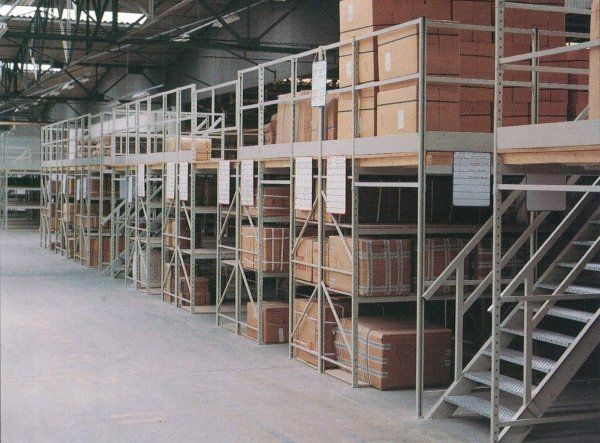 Un magazzino industriale a più livelli con scaffalature metalliche su cui sono impilate scatole marroni, e una scala di accesso.