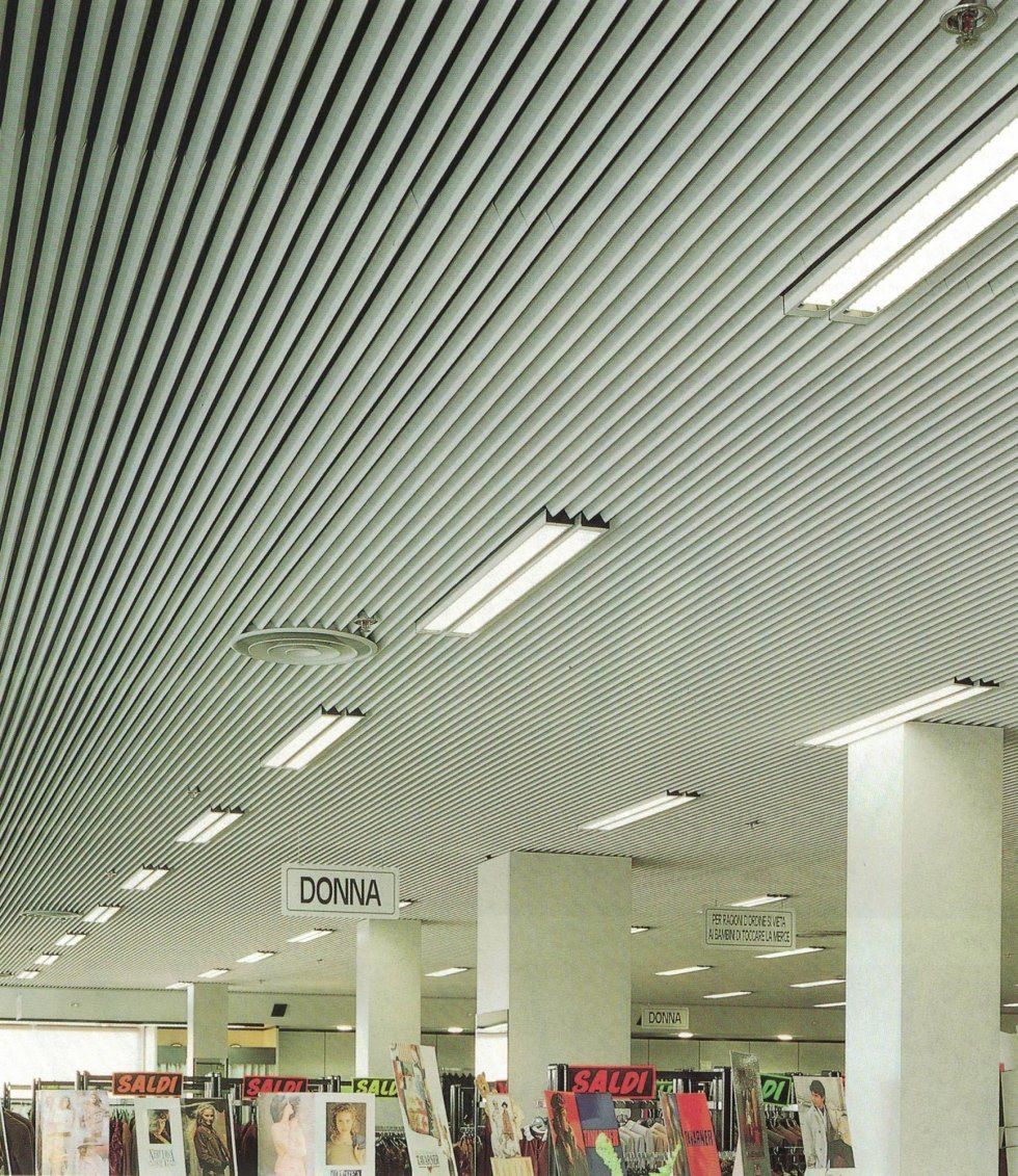 Interno di un negozio al dettaglio caratterizzato da un soffitto a listelli metallici lineari con motivo decorativo, illuminazione a incasso e pilastri di sostegno.