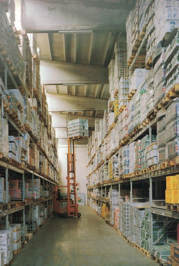 Un carrello elevatore, in uno stretto corridoio di un magazzino, solleva un pallet di merci verso uno scaffale alto tra file di scaffali pieni.