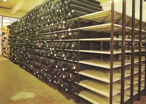 Un grande magazzino con scaffalature industriali stipate di rotoli cilindrici scuri di tessuto, impilati uno sull'altro.