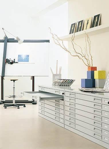 Un ufficio luminoso con un grande tavolo da disegno, uno sgabello e lunghi cassetti in metallo grigio chiaro contenenti cubi colorati.
