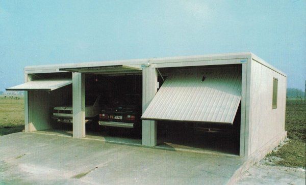 Un garage in cemento a tre vani con portoni basculanti bianchi, che ospita veicoli in un paesaggio pianeggiante e aperto.