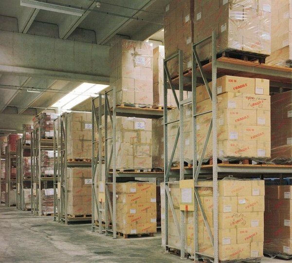 In un magazzino, file di scaffalature industriali metalliche, impilate fino a formare un cumulo di scatole di cartone marrone su pallet di legno.