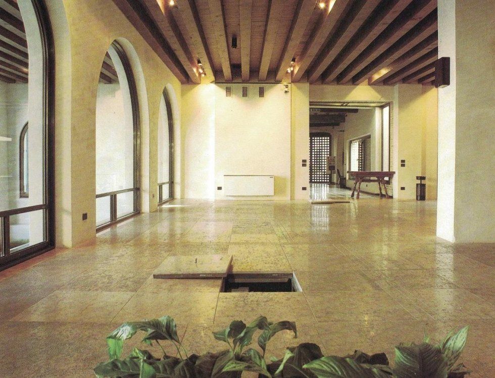 Uno spazio interno con pavimenti in pietra, finestre ad arco e soffitto con travi in legno. Una botola sul pavimento è parzialmente aperta.