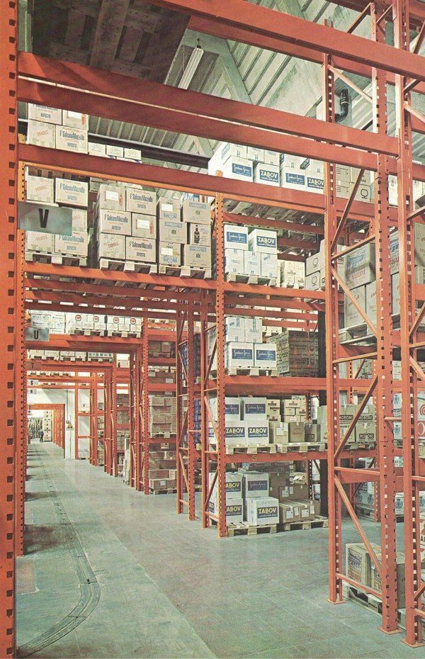 Corridoi di stoccaggio di un magazzino industriale con scaffalature metalliche arancioni piene di scatole impilate su pallet.
