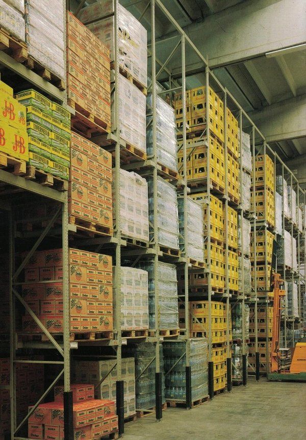 Scaffalature industriali alte e ingombranti, piene di pile di scatole e casse pallettizzate di vari colori.