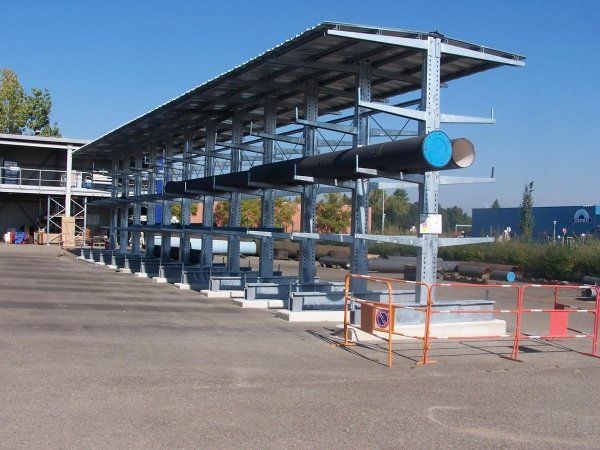 Struttura di stoccaggio industriale in metallo a sbalzo con tettoia, per tubi di grandi dimensioni, da utilizzare all'aperto su un piazzale pavimentato.
