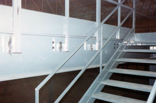 Una scala in metallo con corrimano grigio chiaro e pannelli laterali bianchi, che conduce a una piattaforma rialzata.